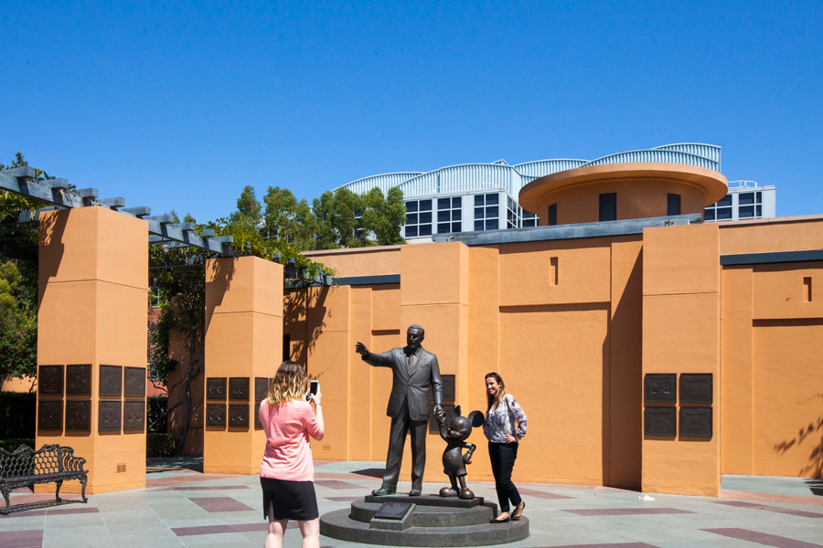 Two women are taking photos with the bronze statue of Walt Disney and Mickey Mouse.