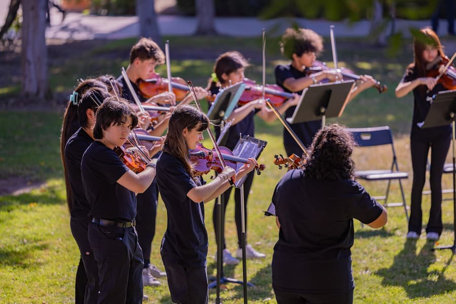 Group of student violinists perform outdoors as part of a youth orchestra during the AME Institute event.