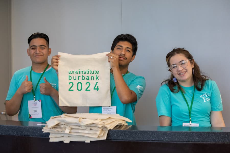 Three volunteers wearing teal shirts stand at a registration table, one holding up a tote bag labeled 'ameinstitute burbank 2024' and smiling.