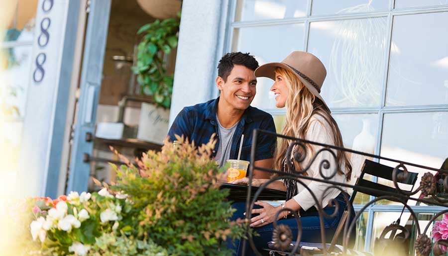 A couple sit together outside at a table while smiling and talking.