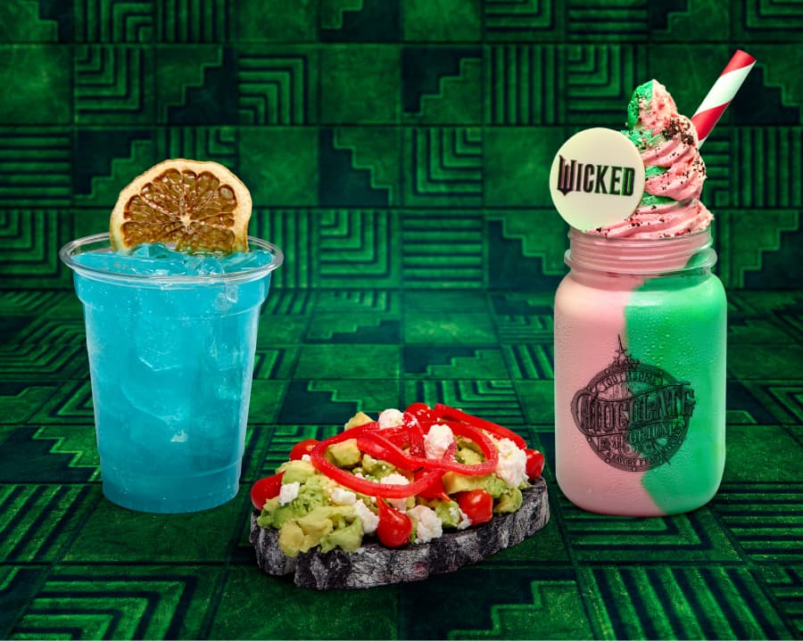 vibrant display of themed food and drinks against a green geometric background. On the left, a clear plastic cup holds a blue icy beverage garnished with a dried lemon slice. In the center, a piece of dark bread topped with mashed avocado, red bell pepper strips, cherry tomatoes, and crumbled feta cheese. On the right, a mason jar filled with a pink and green layered milkshake, topped with whipped cream, colorful sprinkles, and a white chocolate disc featuring the word "Wicked." A red and white striped straw sticks out from the shake.